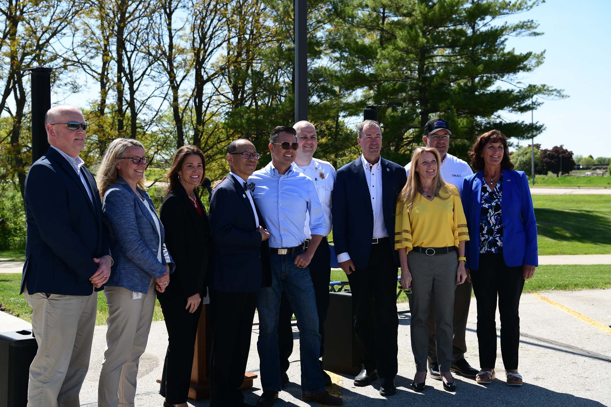 A group of about ten people stand outdoors on a paved area near trees, posing together in business and business-casual attire beside a podium and speakers at a daytime event.