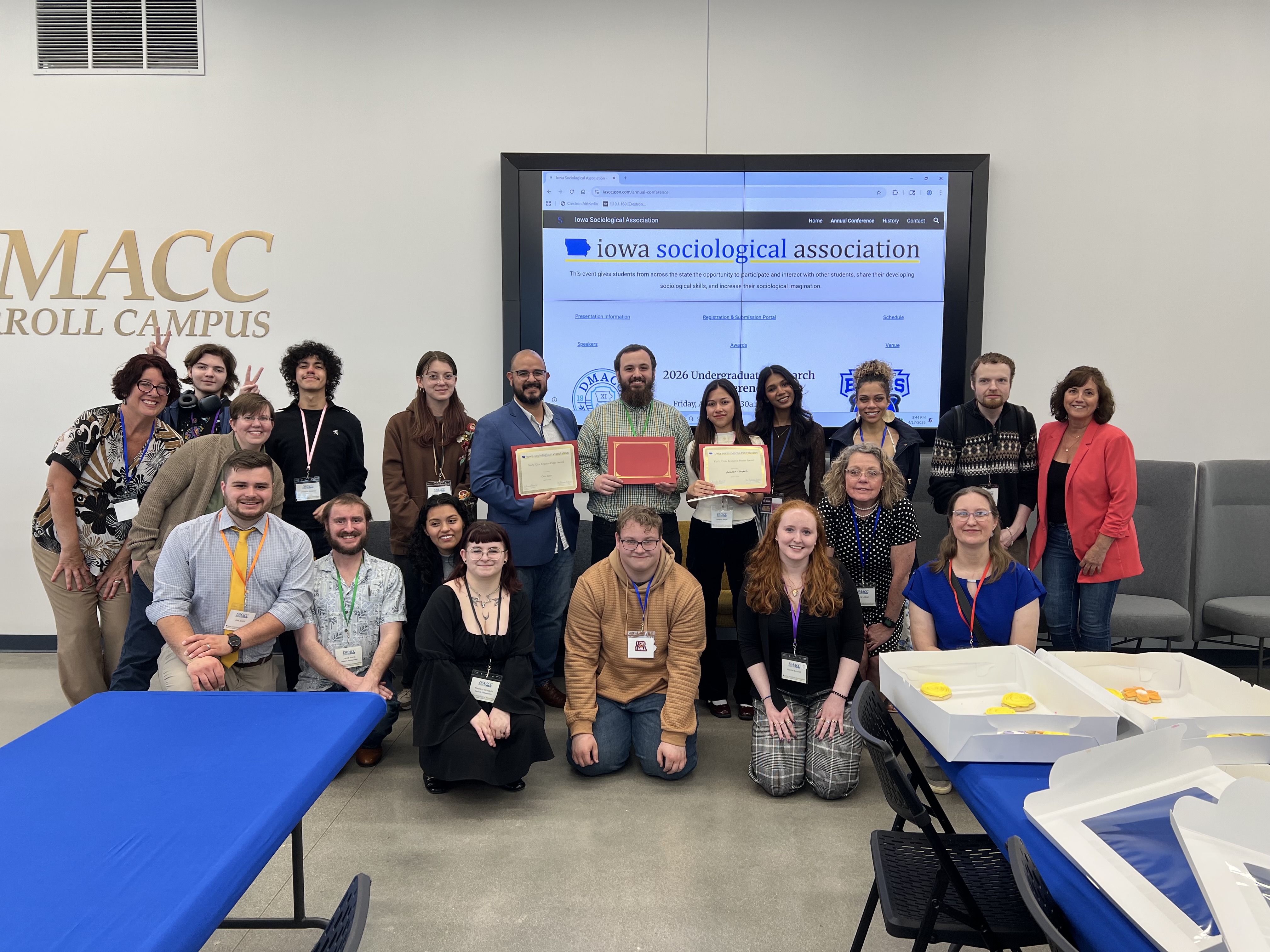 A group of 20 people pose for a photo, the front row on their knees, and the back row standing. A projector screen in the background shows the Iowa Sociological Association website.