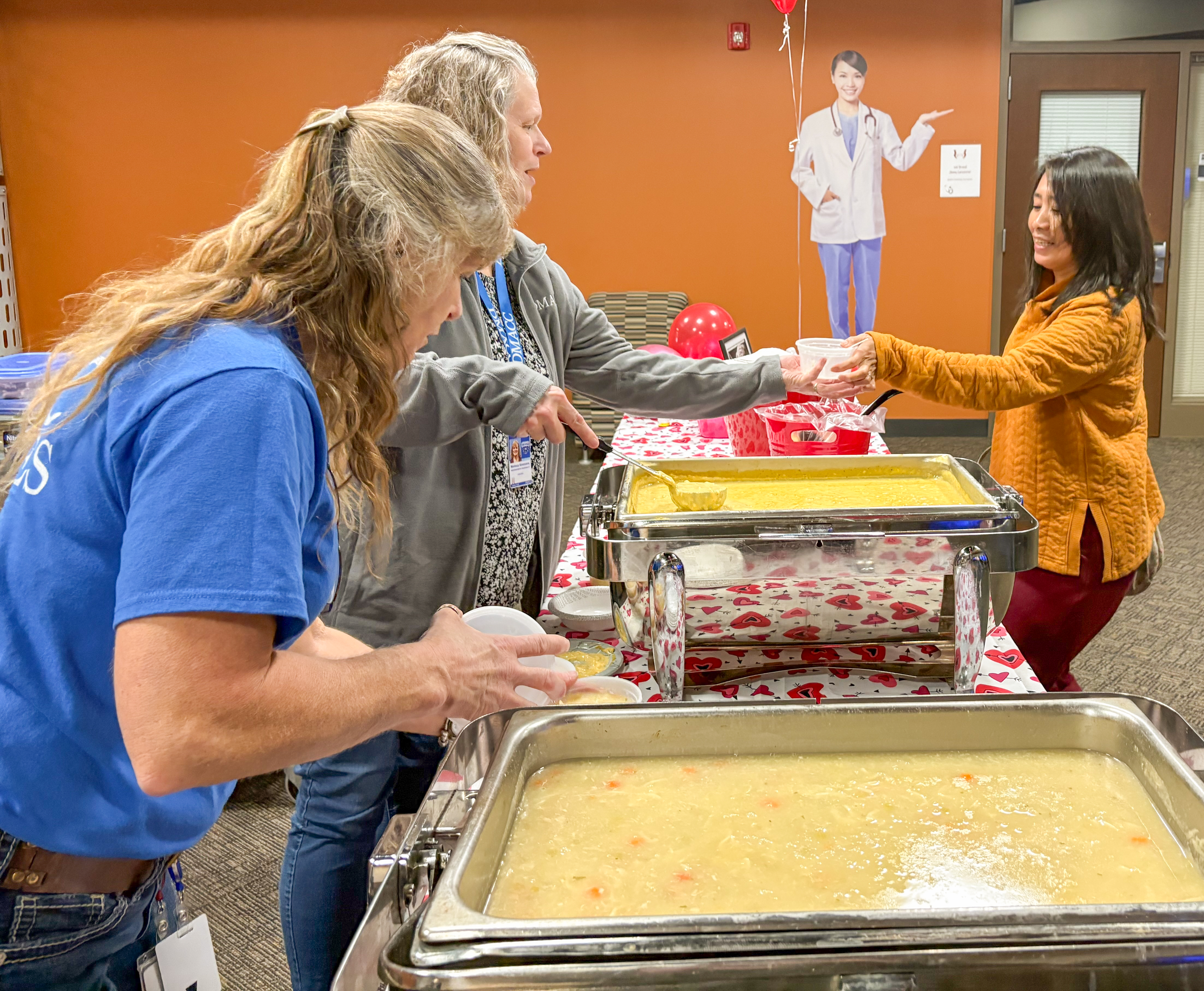Two women in blue shirts serve soup from large pans to a woman in an orange sweatshirt 