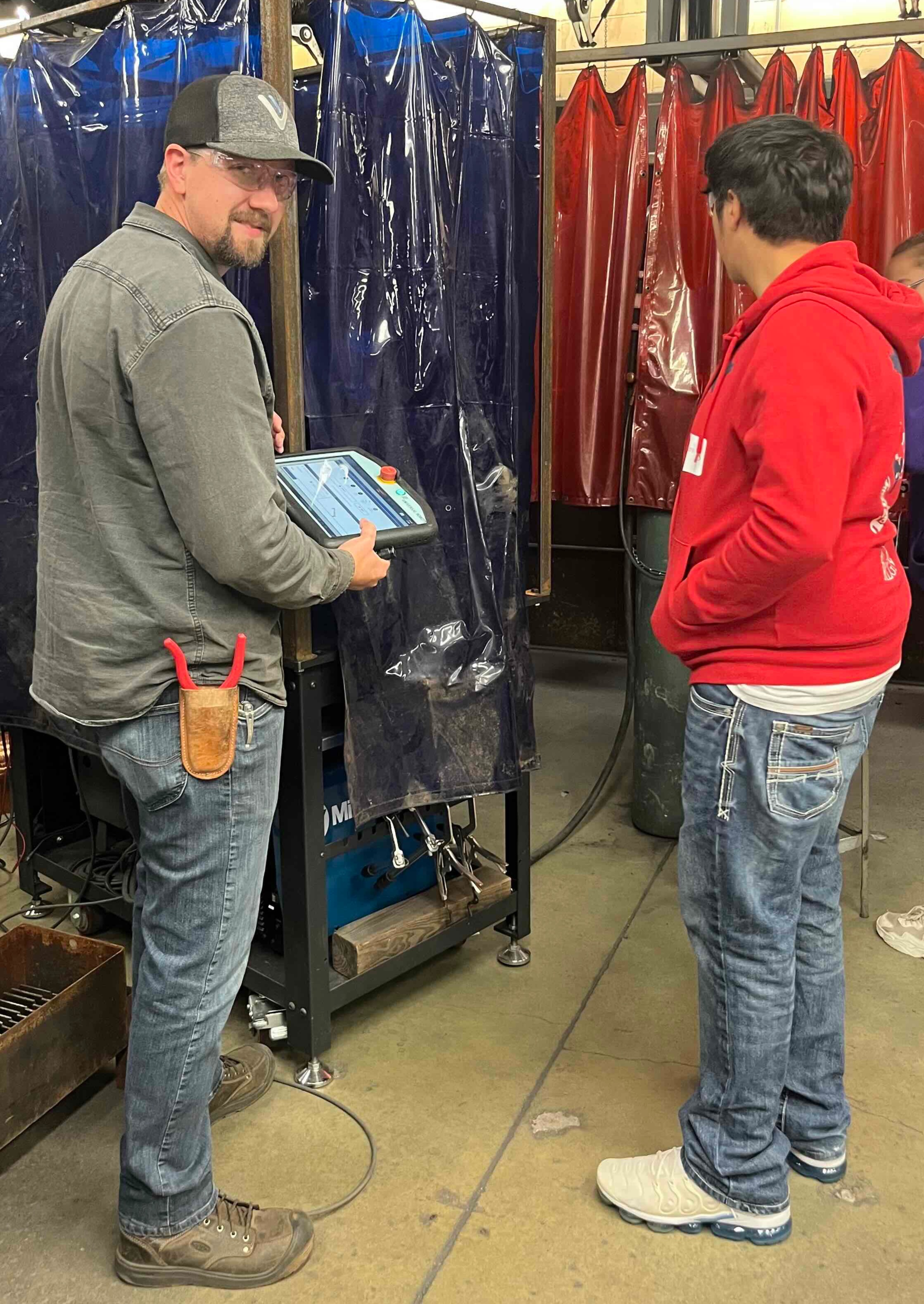 Discover DMACC Manufacturing/Engineering 10/9/26 Standing beside the welding workstation and holding the control interface, Welding Associate Professor Nathan Vander Wert explains the equipment while students observe near enclosed welding booths in the manufacturing lab.