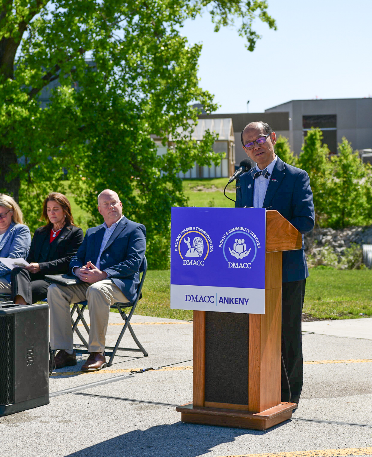 A person speaks at a wooden podium with a microphone and a blue sign reading “DMACC ANKENY” and program icons, while several seated attendees listen in the background on a sunny day.